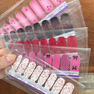 May include: A collection of five Color Street nail polish strip sets in clear plastic packaging. The designs include pink with kiss prints, black with red glitter, solid red glitter, pink with black bows and grid patterns, and white with red and pink glitter.