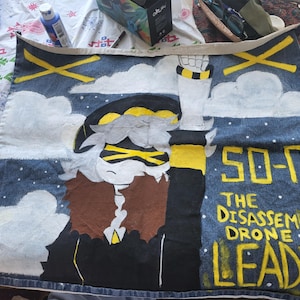 Peut inclure: Tissu en denim peint à la main représentant un personnage de "Murder Drones". L'œuvre comprend un personnage avec des accents jaunes, le texte "SO-U THE DISASSEMBLY DRONE LEADER" et des nuages blancs sur fond bleu.