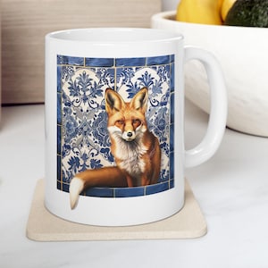 May include: White ceramic mug featuring a detailed illustration of a fox with orange and white fur. The fox is set against a backdrop of blue and white patterned tiles. The mug has a curved handle and sits on a beige coaster.