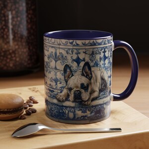 Puede incluir: Taza de cerámica con interior y asa azules, con una ilustración detallada de un bulldog francés descansando sobre una superficie de azulejos azules y blancos. La taza está sobre una superficie de madera con una cuchara y un macarrón.