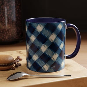 Könnte beinhalten: Eine blau-weiß karierte Kaffeetasse mit dunkelblauem Innenraum und Henkel. Ein silberner Löffel, ein Macaron und Kaffeebohnen liegen auf einer Holzoberfläche neben der Tasse.