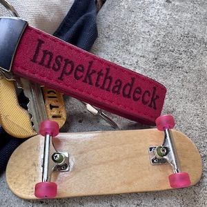 Puede incluir: Un fingerboard con tabla de madera clara, ejes de metal plateado y ruedas rosas translúcidas, colocado debajo de una correa de llavero de tela roja con el texto negro bordado "Inspekthadeck" y un juego de llaves de metal.