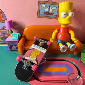 May include: A miniature skateboard with a black deck and pink wheels, featuring a graphic of Homer Simpson. A Bart Simpson figurine sits on an orange couch in a cartoon-themed room. The skateboard has text that reads "How hard I pronounce the F in 'F*** ICE'"