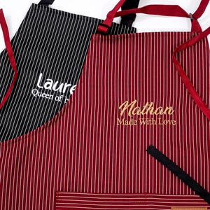 Op de afbeelding: Twee gestreepte schorten. Eén is zwart met witte strepen en de naam "Laure" in wit geborduurd. De andere is bordeauxrood met witte strepen en de naam "Nathan" in goud geborduurd. Beide schorten hebben rode banden.