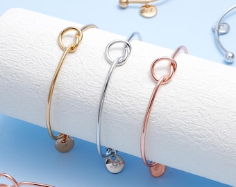 May include: Gold, silver, and rose gold-colored knot bangles. Each bangle features a knot design and a small, round charm. The bangles are displayed on a white cylindrical surface against a light blue background.