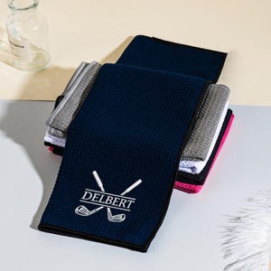 May include: A selection of golf towels in navy blue, grey, white, and pink. The navy towel displays the name "DELBERT" with crossed golf clubs in white. The towels have a textured waffle weave, ideal for absorbing moisture.