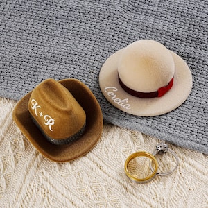 May include: Two miniature hat-shaped ring boxes, one brown cowboy hat and one cream-colored hat, with the names "K+R" and "Cerelia" in white script. A gold and silver ring set rests nearby. The hats are on a textured surface.