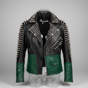 May include: Black and green leather jacket with silver spikes on the shoulders and sleeves. The jacket features a zip closure, a small pocket, and a green lower section. The jacket is displayed on a clear mannequin.