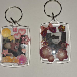 May include: Two clear acrylic keychains with silver key rings. One keychain features a cartoon character with decorative embellishments including stars, hearts, and a flower. The other keychain has a black and white image with a bow and other decorative elements.