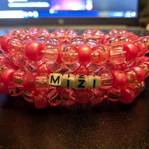 May include: A layered kandi bracelet in shades of red and clear beads. The bracelet features the word "MiZi" spelled out in white letter beads. The beads are strung together to form a thick, textured accessory.