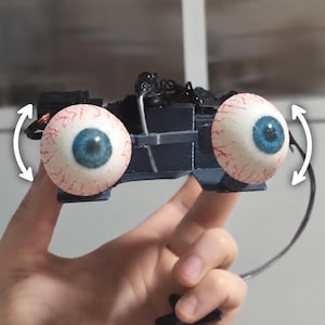 Op de afbeelding: Een mechanisch apparaat met twee realistische, bolvormige oogballen met blauwe irissen en rode aders. De ogen zijn gemonteerd op een donker, hoekig frame met zichtbare draden en kleine motoren, vergezeld van witte gebogen pijlen die beweging aangeven.