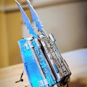 Könnte beinhalten: Ein silberfarbener Unterarmschutz mit zwei spitzen, klingenartigen Vorsprüngen, die nach oben ragen. Das Gerät hat eine komplexe, mechanische Oberfläche mit komplizierten Paneelen, Rippen und einem metallischen Finish, akzentuiert durch ein hellblaues Licht.
