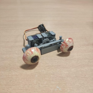 May include: A gray robotic device with two realistic-looking eyeballs as wheels. The eyeballs are white with red veins and brown pupils. The device has a black connector and wires attached.