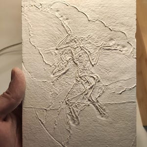 May include: A rectangular off-white plaster plaque featuring a relief carving of a skeletal figure with elongated limbs and a large, cloud-like shape overhead. The surface has a textured, matte finish with visible horizontal lines across the bottom.