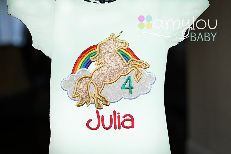 May include: White t-shirt with a gold glitter unicorn, rainbow, and clouds. The number 4 and the name Julia are embroidered on the shirt.