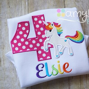 May include: White t-shirt with a pink polka dot number four, a rainbow unicorn, and the name "Elsie" embroidered in rainbow colors. The shirt is folded and has a "amylou BABY" label in the top right corner.
