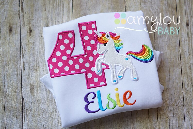 May include: White t-shirt with a pink polka dot number four, a rainbow unicorn, and the name "Elsie" embroidered in rainbow colors. The shirt is folded and has a "amylou BABY" label in the top right corner.