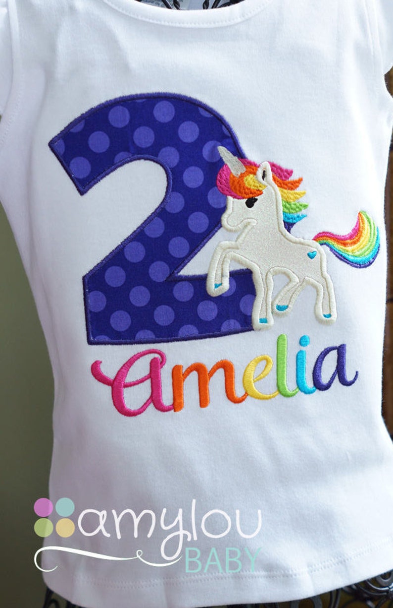 May include: White t-shirt with a purple polka dot number 2 and a rainbow unicorn with the name Amelia embroidered in rainbow colors.