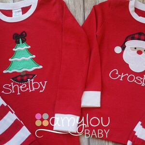 May include: Two red long-sleeved shirts with white striped cuffs. One shirt features a green Christmas tree with a red bow and the name "Shelby". The other shirt features a Santa Claus face with a red and black plaid hat and the name "Crosby".