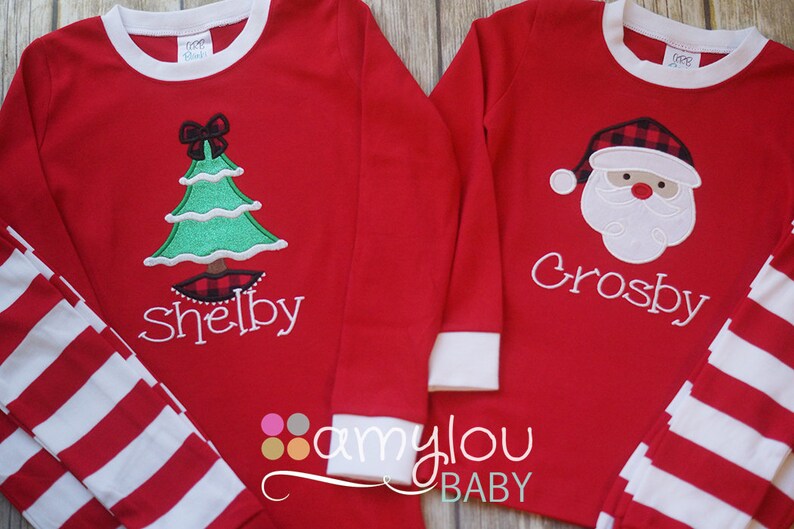 May include: Two red long-sleeved shirts with white striped cuffs. One shirt features a green Christmas tree with a red bow and the name "Shelby". The other shirt features a Santa Claus face with a red and black plaid hat and the name "Crosby".