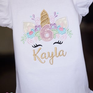 May include: White t-shirt with a gold unicorn design featuring a pink number 6 and the name Kayla embroidered on the front. The shirt has a ruffled hem and the text "amylou baby" is printed on the bottom.