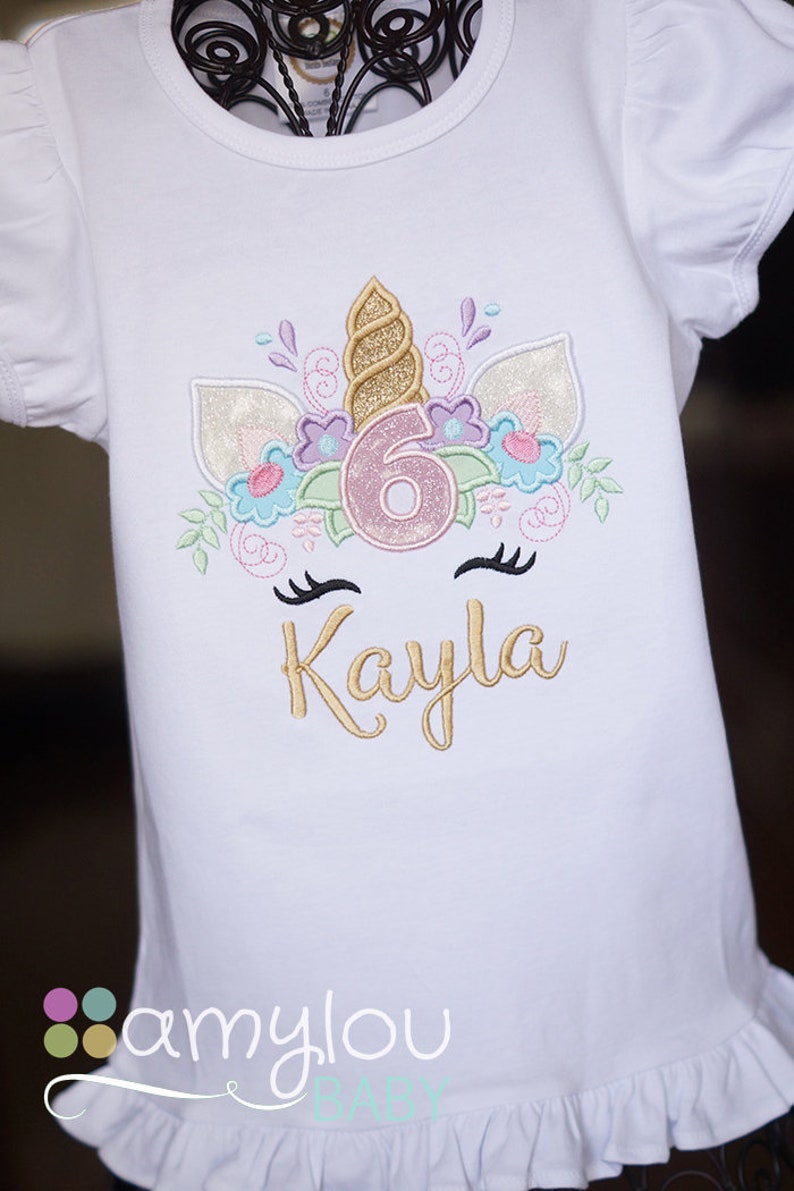 May include: White t-shirt with a gold unicorn design featuring a pink number 6 and the name Kayla embroidered on the front. The shirt has a ruffled hem and the text "amylou baby" is printed on the bottom.