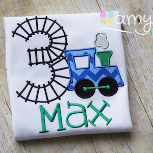 May include: A white t-shirt with a black embroidered number "3" surrounded by a train track design. A blue and green train with a white smoke plume is embroidered to the right of the number. The name "Max" is embroidered in green below the train.
