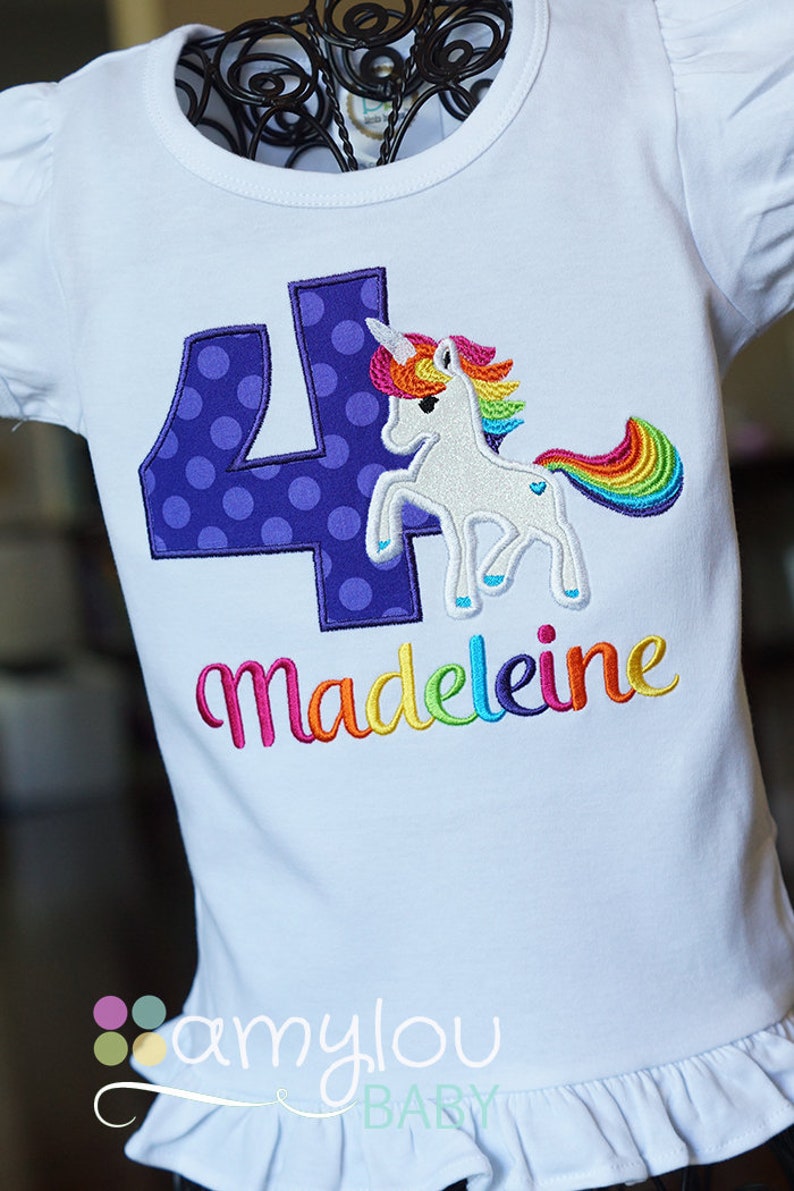 May include: White t-shirt with a purple number four with white polka dots, a rainbow unicorn, and the name "madeleine" in rainbow letters. The shirt has a ruffled hem and the text "amylou baby" at the bottom.