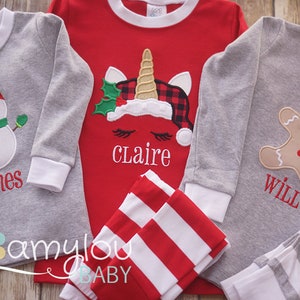 May include: Three pairs of Christmas pajamas for children. The pajamas are gray with white stripes and feature a snowman, a unicorn, and a gingerbread man. The pajamas are personalized with the names "James", "Claire", and "Will".