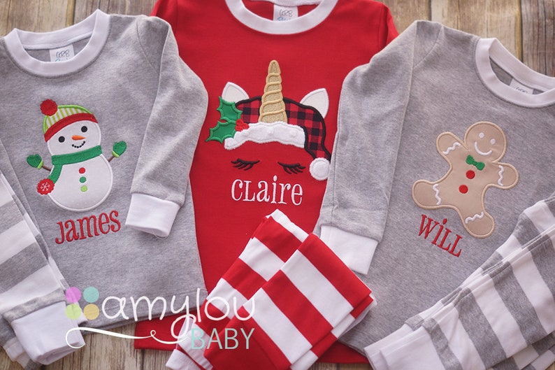 May include: Three pairs of Christmas pajamas for children. The pajamas are gray with white stripes and feature a snowman, a unicorn, and a gingerbread man. The pajamas are personalized with the names "James", "Claire", and "Will".