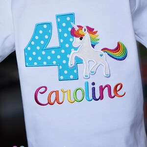 May include: White t-shirt with a blue number four with white polka dots, a rainbow unicorn, and the name "Caroline" embroidered in rainbow colors.