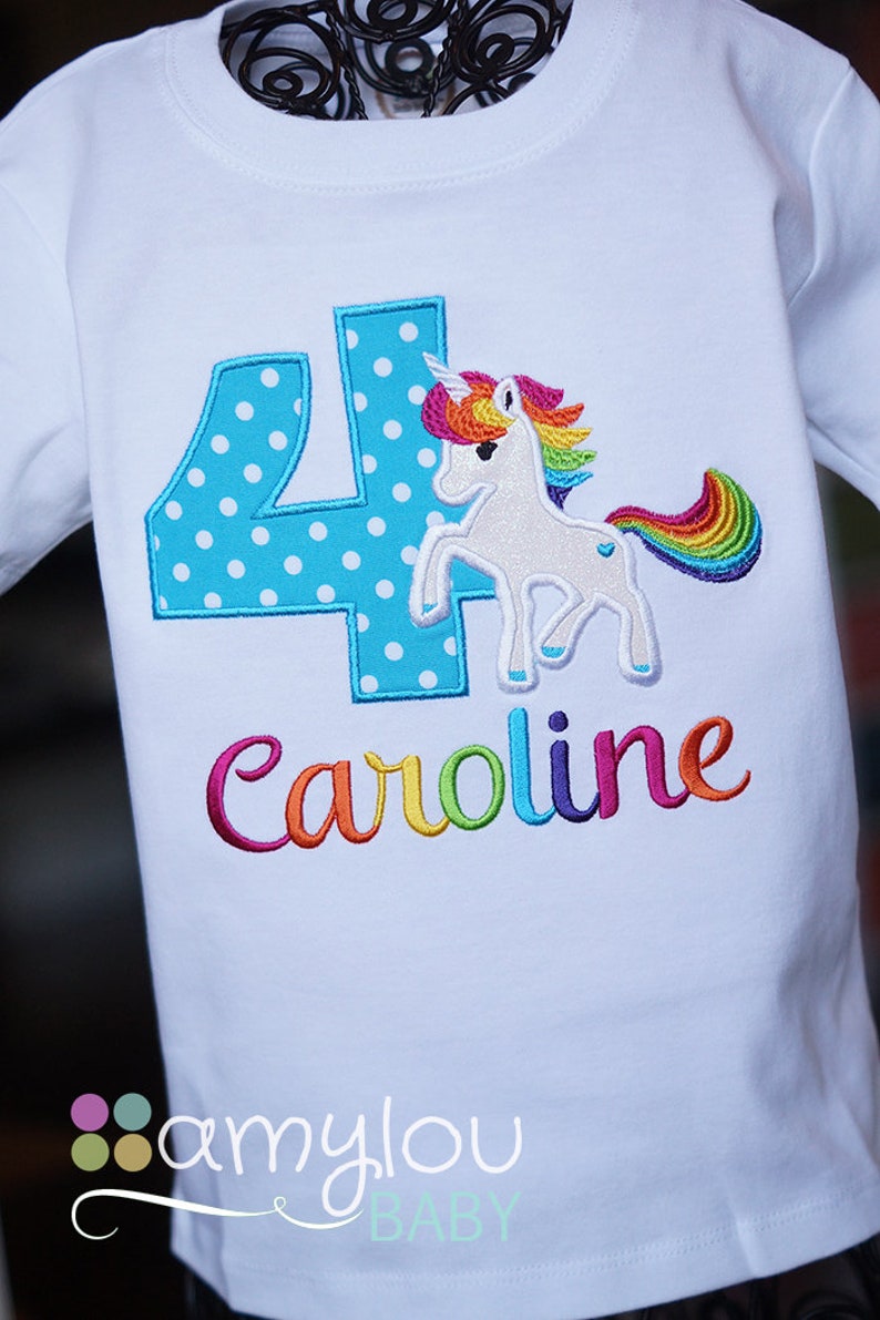 May include: White t-shirt with a blue number four with white polka dots, a rainbow unicorn, and the name "Caroline" embroidered in rainbow colors.
