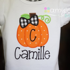 May include: White t-shirt with an orange pumpkin with black and white checkered bow, green stem, and the letter "C" in the center. The name "Camille" is embroidered below the pumpkin.