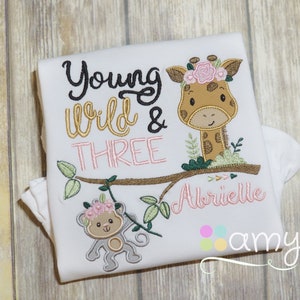 May include: White onesie with embroidered text "Young Wild & Three" and a giraffe and monkey with pink flowers. The name "Abrielle" is embroidered below the giraffe.