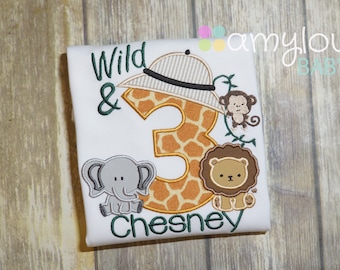 Può includere: Maglietta bianca con ricamo verde che dice "Wild & 3 Chesney". Il numero 3 è una stampa di giraffa con un cappello da safari in cima. Ci sono anche immagini ricamate di un elefante, un leone e una scimmia.