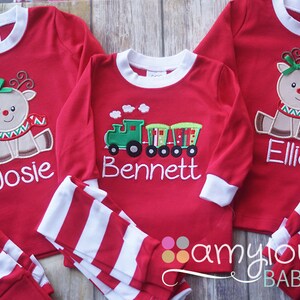 May include: Three red long-sleeved pajamas with white stripes on the sleeves and pants. The pajamas are personalized with embroidered designs and names: Josie, Bennett, and Ellie. The designs include a reindeer, a train, and a reindeer.