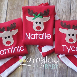 May include: Three red long-sleeved shirts with a reindeer applique and personalized names: Dominic, Natalie, and Tyce. The shirts are folded on top of red and white striped fabric.