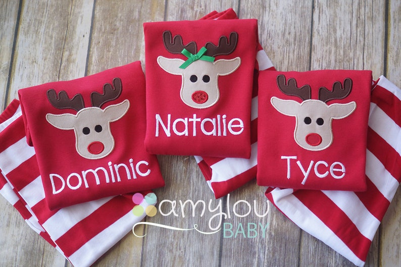 May include: Three red long-sleeved shirts with a reindeer applique and personalized names: Dominic, Natalie, and Tyce. The shirts are folded on top of red and white striped fabric.