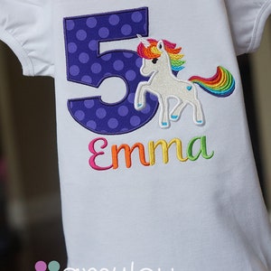 May include: White t-shirt with a purple polka dot number 5 and a rainbow unicorn with the name "Emma" embroidered on it.