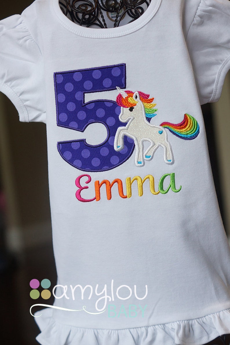 May include: White t-shirt with a purple polka dot number 5 and a rainbow unicorn with the name "Emma" embroidered on it.