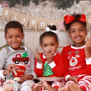 May include: Three children wearing matching Christmas pajamas. The child on the left is wearing a gray pajama top with a red truck and a green tree design. The child in the middle is wearing a red pajama top with a green Christmas tree design. The child on the right is wearing a red pajama top with a reindeer design. The pajamas all have white trim. The children are sitting on a red and white checkered couch with a banner that reads "Silent Night" hanging above them.