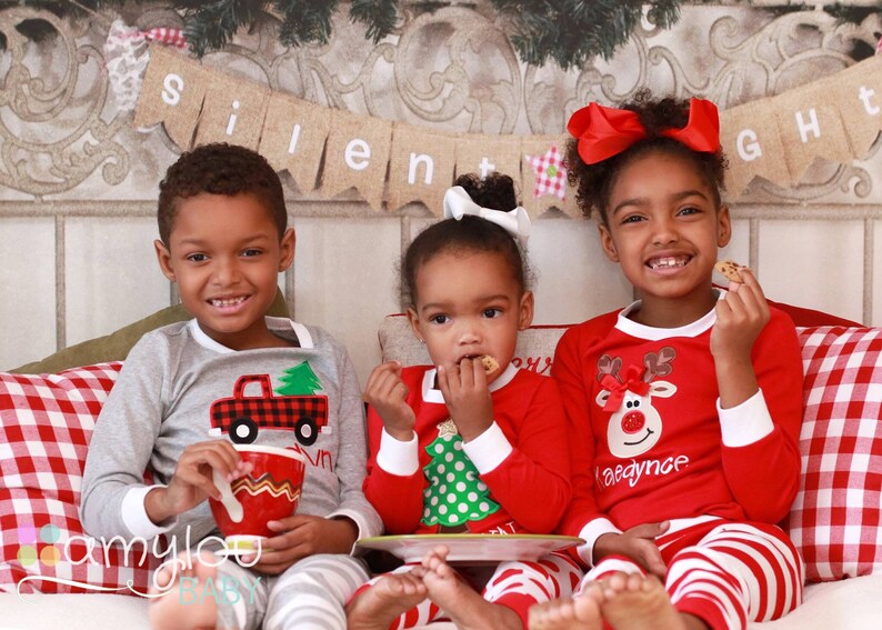 May include: Three children wearing matching Christmas pajamas. The child on the left is wearing a gray pajama top with a red truck and a green tree design. The child in the middle is wearing a red pajama top with a green Christmas tree design. The child on the right is wearing a red pajama top with a reindeer design. The pajamas all have white trim. The children are sitting on a red and white checkered couch with a banner that reads "Silent Night" hanging above them.