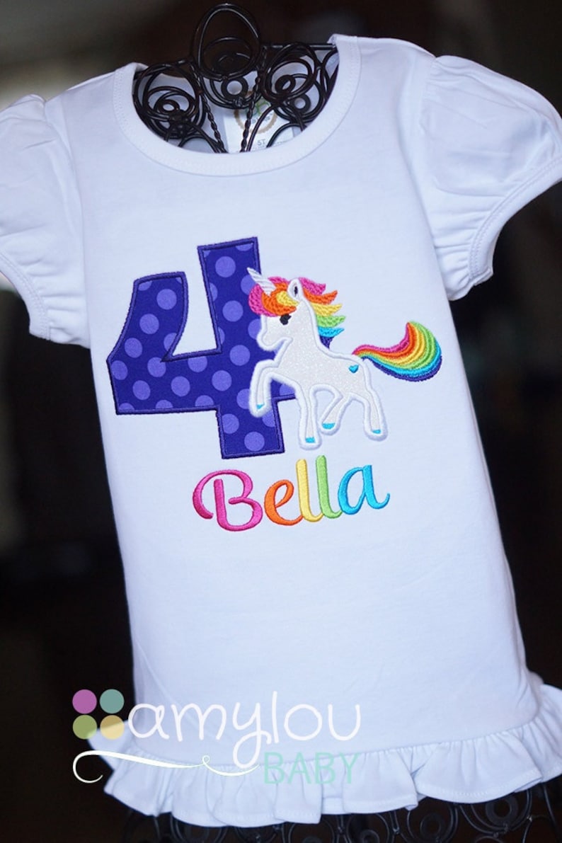 May include: White t-shirt with a purple number four with white polka dots, a rainbow unicorn, and the name "Bella" embroidered in rainbow colors. The shirt has a ruffled hem.