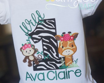 May include: White t-shirt with a green "wild" text, a zebra-patterned number one, a monkey and a giraffe with flowers, and the name "Ava Claire" in green text.