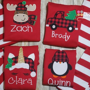 May include: Four red and white striped shirts with different embroidered designs. The designs include a moose, a truck with a Christmas tree, a unicorn, and a penguin. Each shirt has a name embroidered on it: Zach, Brody, Clara, and Quinn.