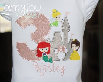 May include: A white t-shirt with a pink glittery number 3, a silver glittery castle, a pink bow, a mermaid, a princess, and a fairy. The text "Karley" is embroidered in pink below the design.