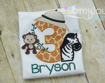 Può includere: Maglietta bianca con un ricamo a tema safari. Il ricamo presenta un numero "3" con stampa di giraffa, una scimmia, una zebra e un cappello da safari. Il nome "Bryson" è ricamato sotto il disegno.