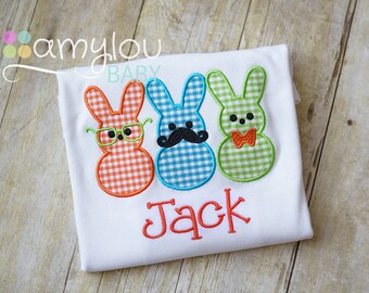 May include: White t-shirt featuring three gingham bunny appliqués in orange, blue, and green, each with unique accessories: glasses, a mustache, and a bow tie. The name "Jack" is embroidered in orange script below the bunnies.