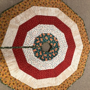 May include: A colorful Christmas tree skirt with a circular design. The skirt features concentric rings of red, white, green, and gold fabrics. The center ring is green with gingerbread men. The outer edge has a leaf pattern.