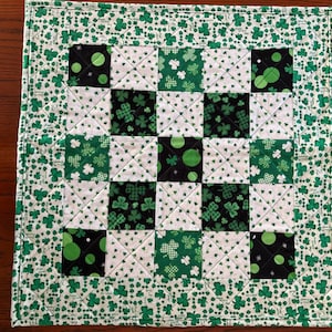 May include: A square quilted placemat with a St. Patrick's Day theme. The center features a checkerboard pattern of black, white, and green squares with shamrock and polka dot designs. The border is white with a repeating pattern of green shamrocks.